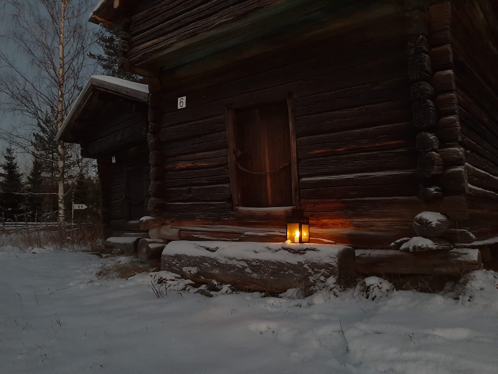 Pienen vanhan hirsirakennuksen portaalla palaa kynttilä lyhdyssä