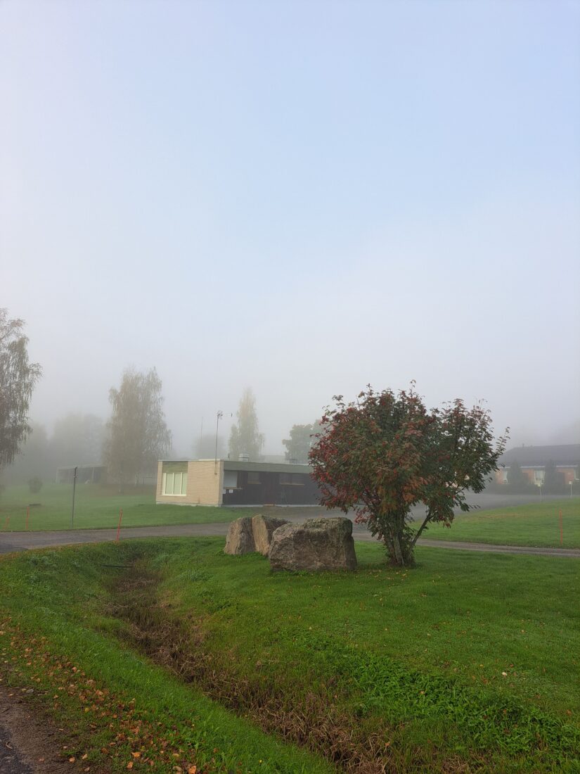 Sumuisessa maisemassa näkyy matala kulmikas huoltorakennus, jonka seinässä on kunnan vesipiste.