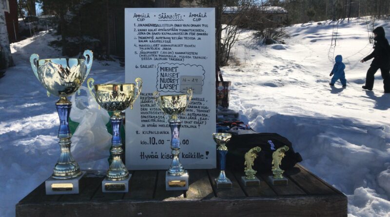 Pöydällä on muutama eri kokoinen palkintopysti ja niiden takana nojallaan valkoinen taulu, johon kirjoitettu pilkkikilpailun säännöt.